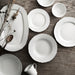 IKEA UPPLAGA tableware collection, including an oval serving platter, plates, bowls, and a small cup, arranged on a wooden surface with a wooden spoon and dried plants—elegant white ceramic set for refined dining -60438022.