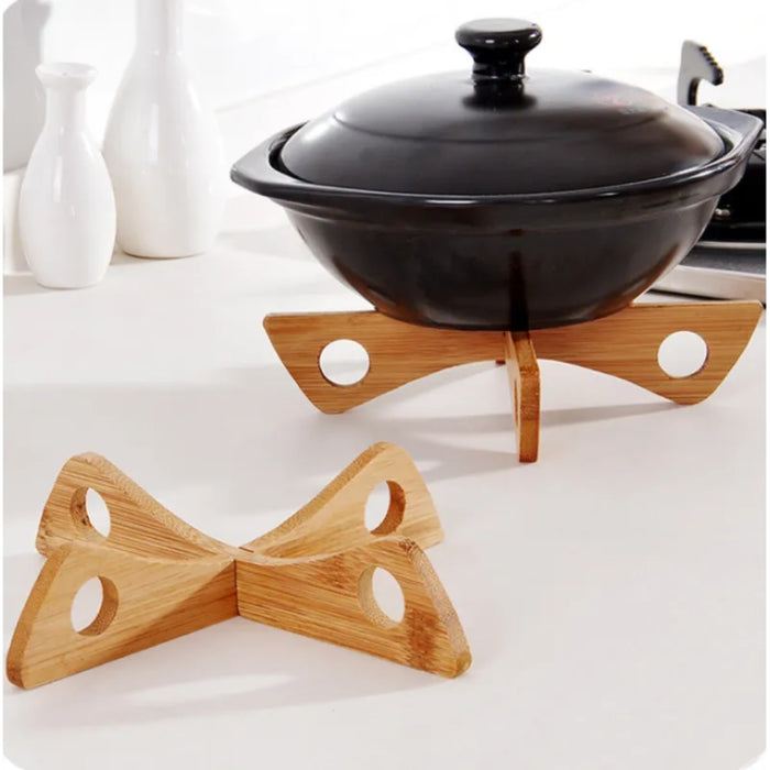 Black ceramic pot with lid resting on a uniquely designed wooden trivet with four triangular legs, each featuring a circular cutout; another identical trivet lies in front for display. Background elements include two sleek white ceramic bottles—likely for oil or vinegar—and part of a stovetop, creating a cozy, modern kitchen vibe – 562152 12826862.