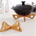Black ceramic pot with lid resting on a uniquely designed wooden trivet with four triangular legs, each featuring a circular cutout; another identical trivet lies in front for display. Background elements include two sleek white ceramic bottles—likely for oil or vinegar—and part of a stovetop, creating a cozy, modern kitchen vibe – 562152 12826862.
