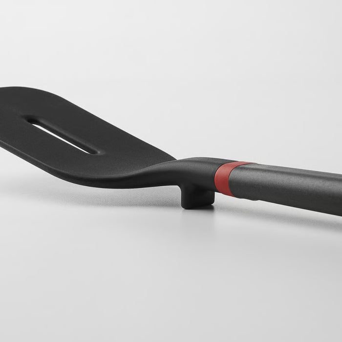 A modern black spatula with a flat head and a single slot, featuring a gray handle with a red accent and a built-in rest that keeps the head elevated off the surface.