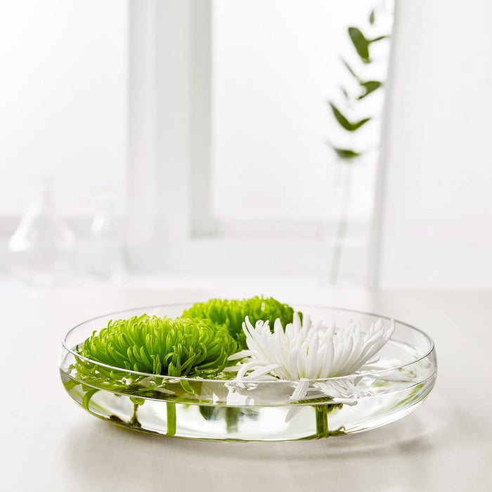 IKEA BERÄKNA bowl—clear glass, shallow design (26 cm diameter), filled with water and three floating flowers (two green, one white); set in a serene scene with soft light through a window, leafy plant in the background, and scattered lab glassware for a minimalist botanical touch – 80329041.