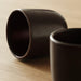 Two dark reddish-brown ceramic mugs with subtle speckles on a light wooden surface. One mug is lying on its side, showing the textured rim and dark interior, while the other is upright and slightly out of focus-80592418