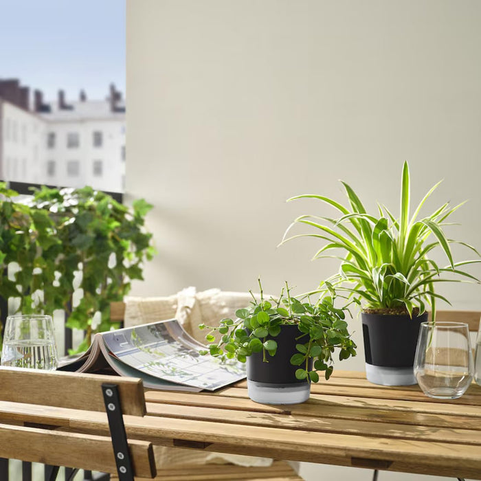 IKEA VÅRDTRÄD self-watering plant pot in transparent with anthracite base—placed on a sunny balcony with leafy plants, an open magazine, and a glass of water for a relaxing greenery vibe – 80570869.