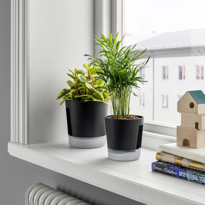 IKEA VÅRDTRÄD self-watering plant pots on a bright windowsill—two pots with transparent tops and anthracite bases hold leafy indoor plants; one with variegated green-yellow leaves, the other with slender palm-like foliage, placed beside stacked books and a small wooden house décor – 80570869.