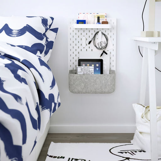 A neatly organized bedside space featuring an IKEA SKÅDIS pegboard with a white shelf and felt pocket. The setup includes a digital clock, a book, a small box, nail polish, and hanging headphones.-60599519