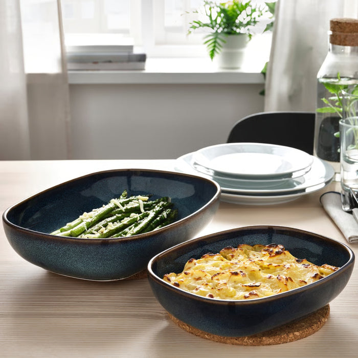 Elegant table setting featuring two IKEA GLADELIG blue ceramic oven dishes—one filled with asparagus spears garnished with grated cheese, the other with a baked potato gratin displaying a golden crust. The dishes are placed on a wooden dining table accompanied by white plates, a glass of water, cutlery on a napkin, and decorative elements like a potted plant, a jar with a cork lid, and stacked books on a windowsill—exuding warmth and home-style refinement -20531079.