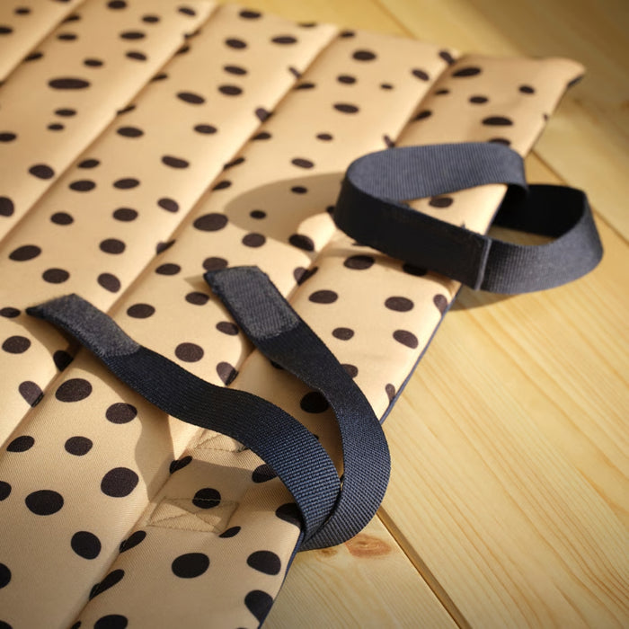 Close-up of beige SANDLÖPARE mat with black polka dots and straps, placed on wooden floor with sunlight highlighting texture