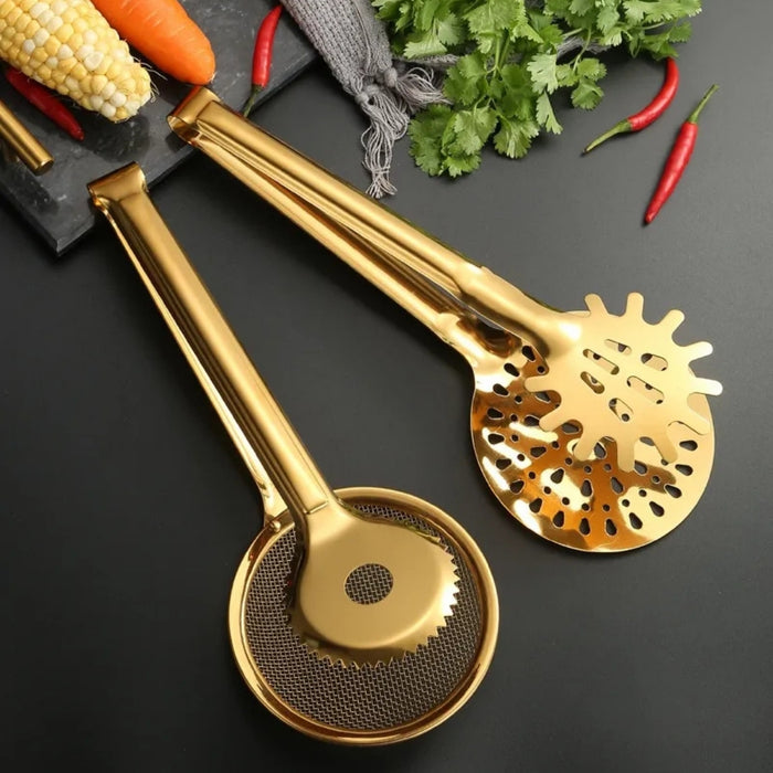 Two golden stainless steel kitchen tongs resting on a dark surface—one designed with a circular mesh strainer and serrated rim, the other featuring an oblong slotted skimmer head with a mix of elongated and round holes. The background includes vibrant cooking elements like corn on the cob, carrots, red chilies, cilantro, and a gray kitchen cloth placed on a black marble board—highlighting the tools' elegant utility in everyday cooking and serving -1600003112383 13034147.
