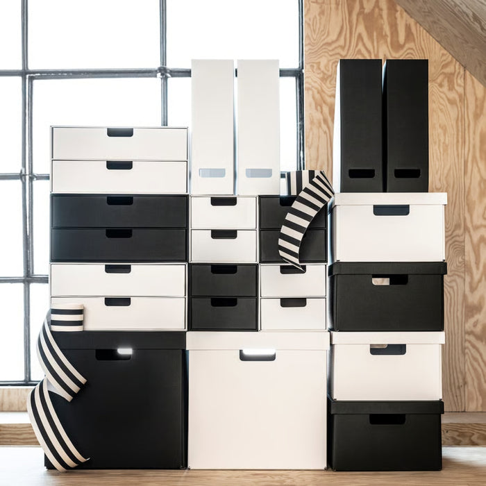 Stack of black and white storage boxes and drawers in a room with wooden floor and window.