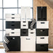 Stack of black and white storage boxes and drawers in a room with wooden floor and window.