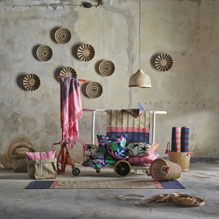 Rustic interior with woven wall hangings, pendant lamp, and MÄVINN throws draped over cart surrounded by colorful cushions and rugs