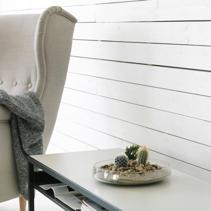 IKEA BERÄKNA bowl—clear glass, 26 cm diameter, shallow design used as a decorative succulent terrarium. Display includes white sand, smooth pebbles, and a mix of small cacti, set atop a white table with black metal legs beside a tufted gray armchair and knit throw. Background features white wooden paneling for a clean, Scandinavian aesthetic – 80329041.