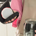 Person using a black jar opener tool to unscrew the metal lid of a glass jar. The user, dressed in a pink shirt and apron, grips the ergonomic handle of the opener firmly positioned on the jar’s lid. Scene set in a home kitchen, highlighting ease-of-use for individuals with limited grip strength or dexterity challenges.- 1598452898401 13017586
