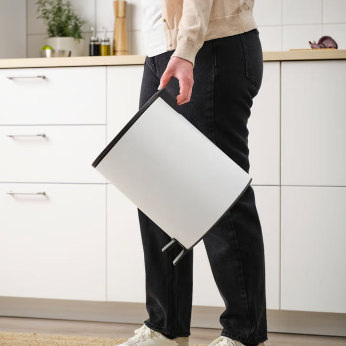 Lifestyle shot of a person carrying the pedal bin in a kitchen with white cabinets.