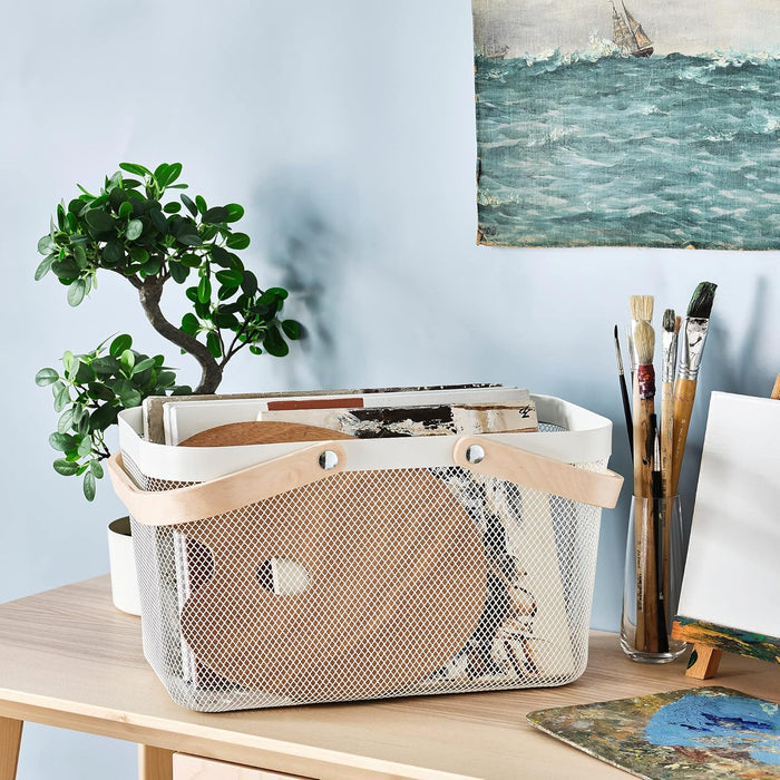 An IKEA RISATORP white wire mesh basket with a wooden handle, filled with art supplies like sketchbooks, a wooden palette, and other flat items, sitting on a light wooden desk next to a plant and paintbrushes in a glass.-90530397