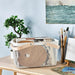 An IKEA RISATORP white wire mesh basket with a wooden handle, filled with art supplies like sketchbooks, a wooden palette, and other flat items, sitting on a light wooden desk next to a plant and paintbrushes in a glass.-90530397