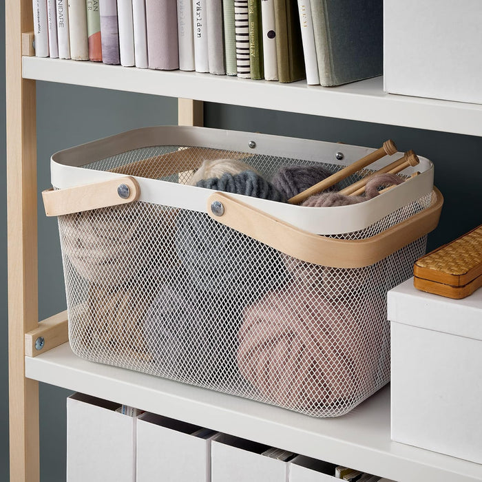 KEA RISATORP white wire mesh basket with a wooden handle, filled with skeins of yarn and knitting needles, sitting on a white shelf below a row of books-90530397