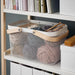 KEA RISATORP white wire mesh basket with a wooden handle, filled with skeins of yarn and knitting needles, sitting on a white shelf below a row of books-90530397