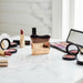 Gold-handled makeup brush surrounded by lipsticks, blushes, and eyeshadow palette lifestyle setting

