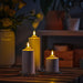 Three SMÅLOM candles of varying heights lit on table with floral vase and shelf decor, creating serene atmosphere