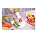 Vegetable peeler being used to peel carrots and celery on a white plate with a kitchen setting in the background.