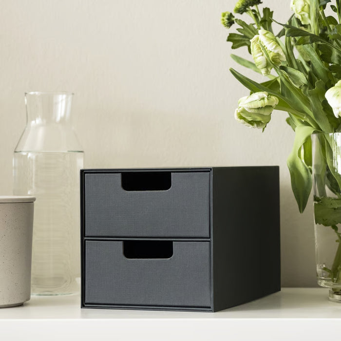 Black two-drawer storage unit on a white surface with a vase and flowers in the background.