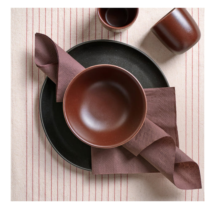 Brown ceramic bowl on a black plate with a folded brown napkin on a striped tablecloth.