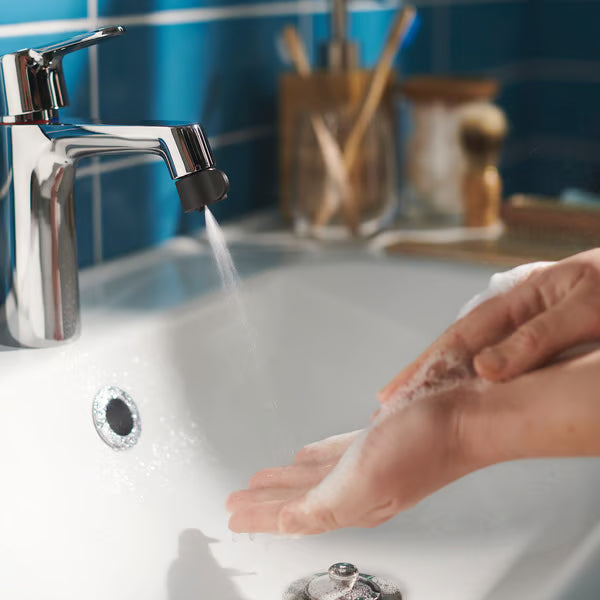 Person washing toothbrush under chrome mixer tap with ÅBÄCKEN nozzle blue-tiled bathroom