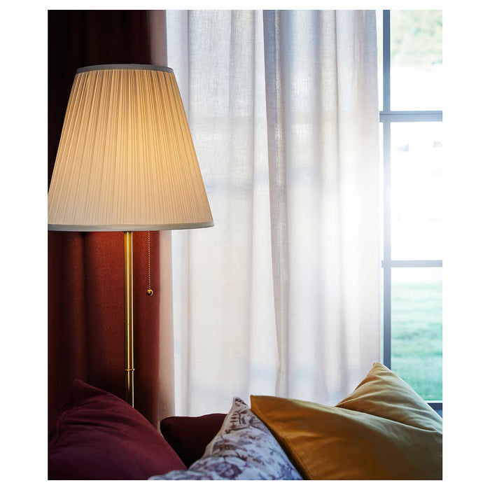 IKEA ÅRSTID brass floor lamp next to window diffusing warm light with red and mustard cushions nearby