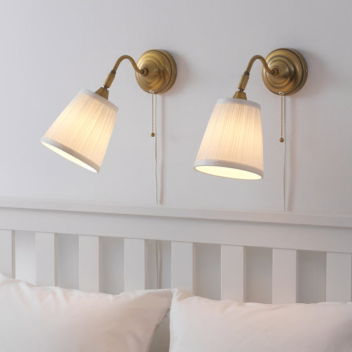  Pair of IKEA ÅRSTID brass wall lamps with pull switches above bed with pink cushions close-up