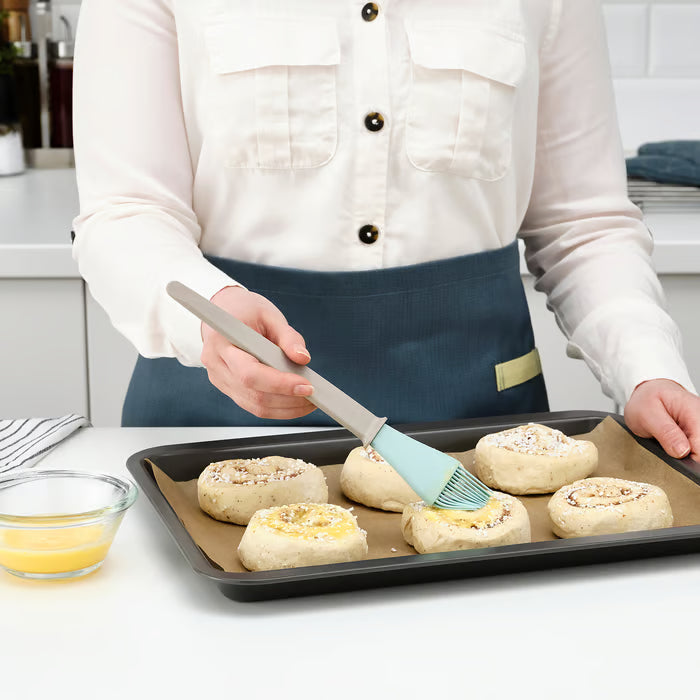 Person in apron using IKEA BAKGLAD blue pastry brush to spread glaze on pastries on a baking tray