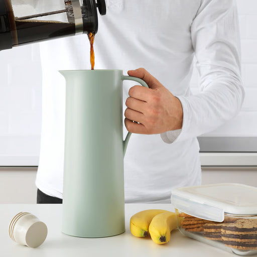 Person filling IKEA BEHÖVD light green vacuum flask on table with bananas and biscuits