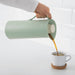 Hand pouring from IKEA BEHÖVD green vacuum flask into white mug showing pour-without-removing-lid feature