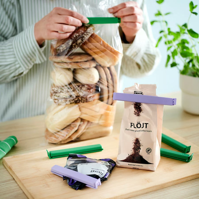 : Person sealing a bread bag with green IKEA BEVARA clips beside coffee bag on wooden table