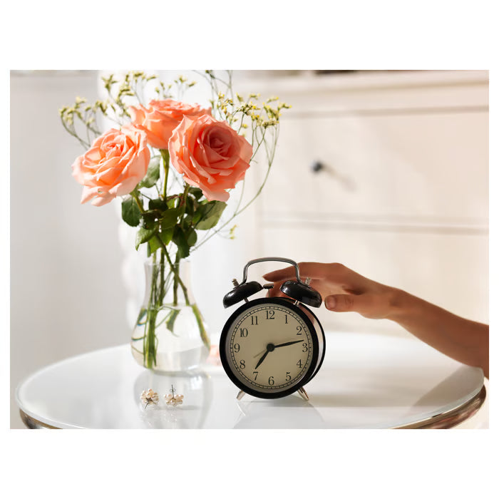 Person setting IKEA DEKAD black analogue alarm clock on white table beside vase with orange roses