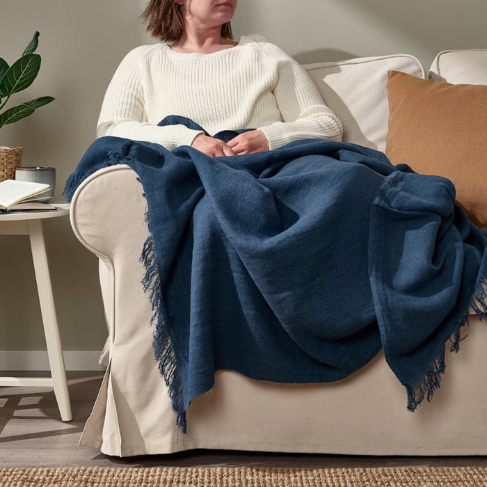Woman comfortably seated on a couch, wrapped in the soft dark blue IKEA DYTÅG throw blanket