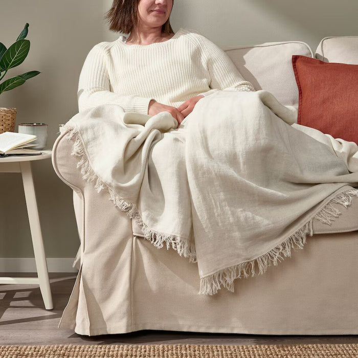 "Woman comfortably seated on a couch, enjoying the soft IKEA DYTÅG off-white throw blanket"