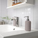 IKEA EKOLN beige soap dispenser on white bathroom counter with modern silver tap, lifestyle setting