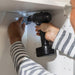 A person is using a cordless drill to install a metal bracket inside a white cabinet. The person is wearing a striped long-sleeve shirt and is holding the drill with one hand while positioning the bracket with the other hand. The cabinet has pre-drilled holes on the side panel-50568085