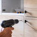 A person is using a black cordless drill to make a hole in a light wood cabinet door. The cabinet has black handles, and there are some tools and parts inside the cabinet-