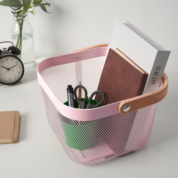 Pink IKEA RISATORP basket, 25x26x18 cm (9 ¾x10 ¼x7 "), placed on a desk with office essentials. Features ventilated steel mesh with a sturdy wooden handle for easy portability. The basket contains books, a green cup with pens, pencils, and scissors, creating a well-organized workspace.-60588691