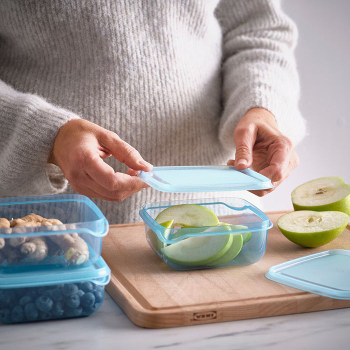 A person handling an IKEA PRUTA food container with a blue lid, 0.6 l (20 oz), filled with green apple slices. Two other containers with ginger and blueberries are stacked nearby on a wooden cutting board, alongside green apple halves-60597105 