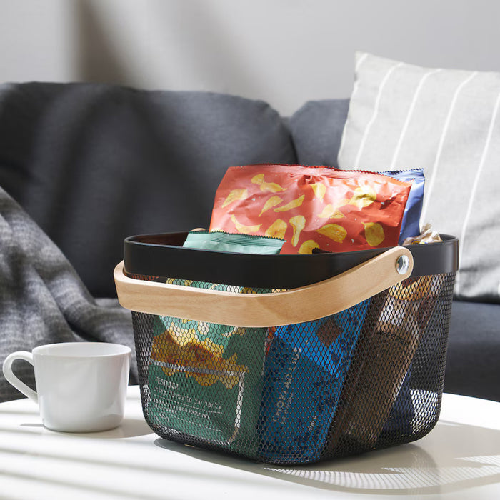 Dark grey IKEA RISATORP basket, 25x26x18 cm (9 ¾x10 ¼x7 "), placed on a desk with organized essentials. Features ventilated steel mesh with a sturdy wooden handle for easy portability. The basket contains books, a green cup holding pens, pencils, and scissors, creating a stylish and functional workspace.-00588712