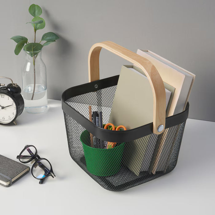 Dark grey IKEA RISATORP basket, 25x26x18 cm (9 ¾x10 ¼x7 "), placed on a desk with office essentials. Features ventilated steel mesh with a sturdy wooden handle for easy portability. The basket contains books, a green cup with pens, pencils, and scissors, creating a well-organized workspace.-00588712