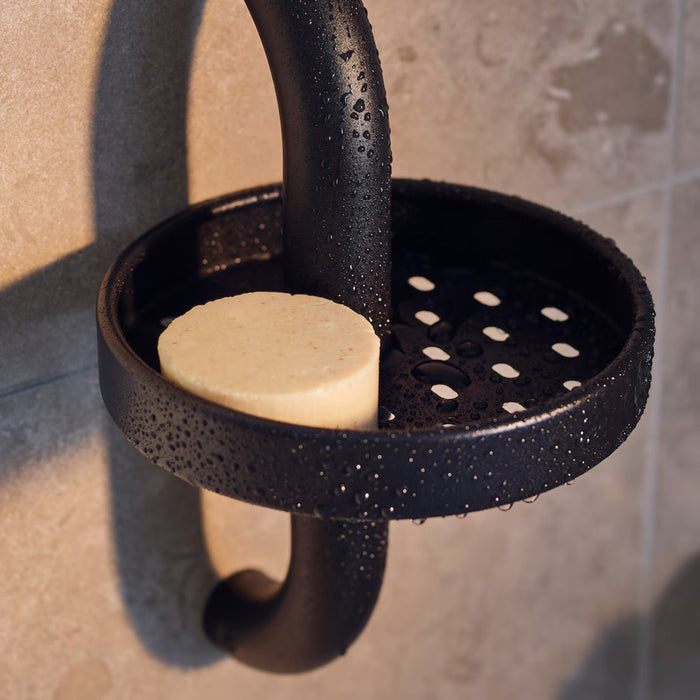 A black shower caddy with a perforated bottom is mounted on a tiled wall. The caddy is wet with visible water droplets and holds a round, slightly used beige bar of soap.-90595992