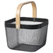 Dark grey IKEA RISATORP basket, 25x26x18 cm (9 ¾x10 ¼x7 "), placed on a white table beside a ceramic mug. Ventilated steel mesh with a sturdy wooden handle for easy portability.-00588712