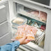 A hand placing an IKEA ISTAD resealable bag with a pale pink geometric pattern into an open freezer drawer. The bag contains food items, possibly pancakes or flatbreads-80592710