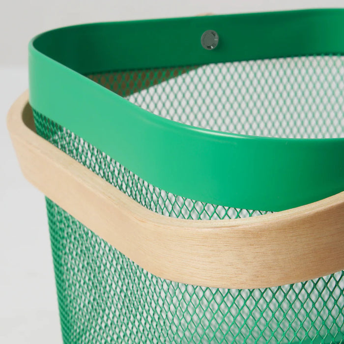 Dark green IKEA RISATORP basket, 25x26x18 cm (9 ¾x10 ¼x7 "), filled with assorted packaged snacks, placed on a white surface beside a ceramic cup. Ventilated steel mesh with a sturdy wooden handle for easy portability. Ideal for stylish and functional kitchen, office, or living room storage. Background features a cozy couch with a gray blanket and a striped pillow, enhancing the inviting aesthetic.- 00588689