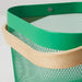 Dark green IKEA RISATORP basket, 25x26x18 cm (9 ¾x10 ¼x7 "), filled with assorted packaged snacks, placed on a white surface beside a ceramic cup. Ventilated steel mesh with a sturdy wooden handle for easy portability. Ideal for stylish and functional kitchen, office, or living room storage. Background features a cozy couch with a gray blanket and a striped pillow, enhancing the inviting aesthetic.- 00588689