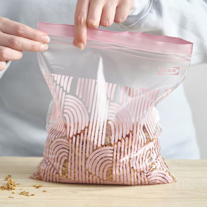 A person placing a cinnamon roll into an IKEA ISTAD resealable bag with a pale pink geometric pattern. The bag features a double-seal strip for secure closure.-80592710